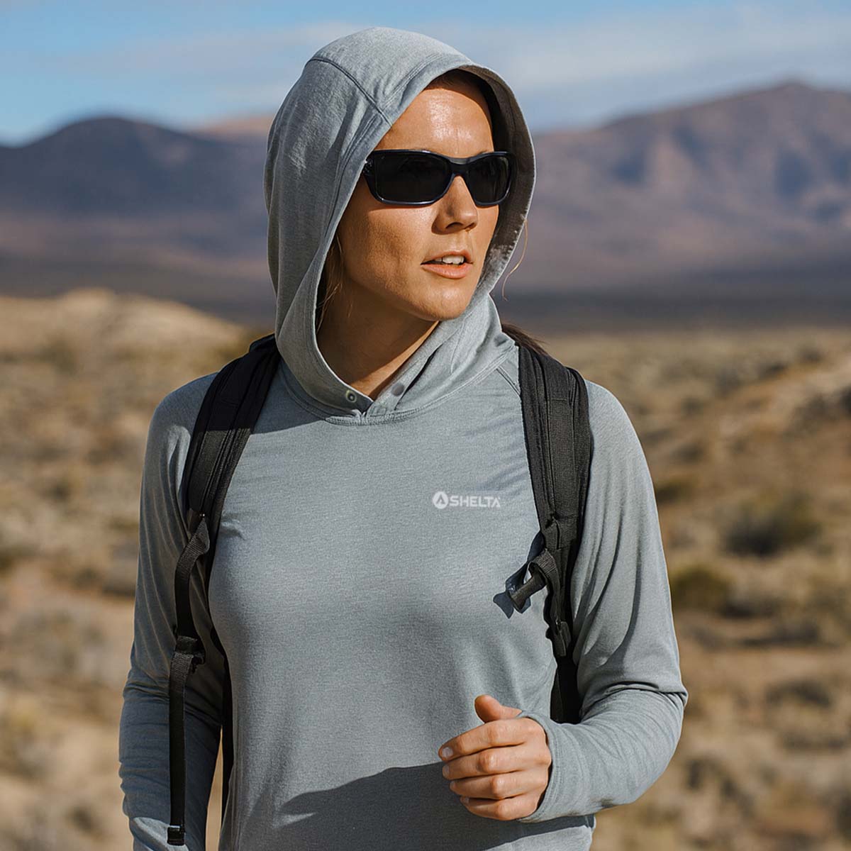 Person wearing a gray hoodie with a brand logo, sunglasses, and a backpack in a desert landscape.