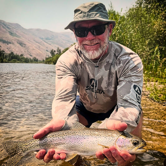 Man holding a fish by a lake with mountains in the background wearing a shelta sun hat