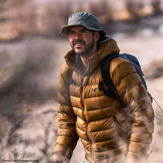 Man wearing a brown puffer jacket and green cap with a backpack, standing in a natural setting wearing a shelta sun hat