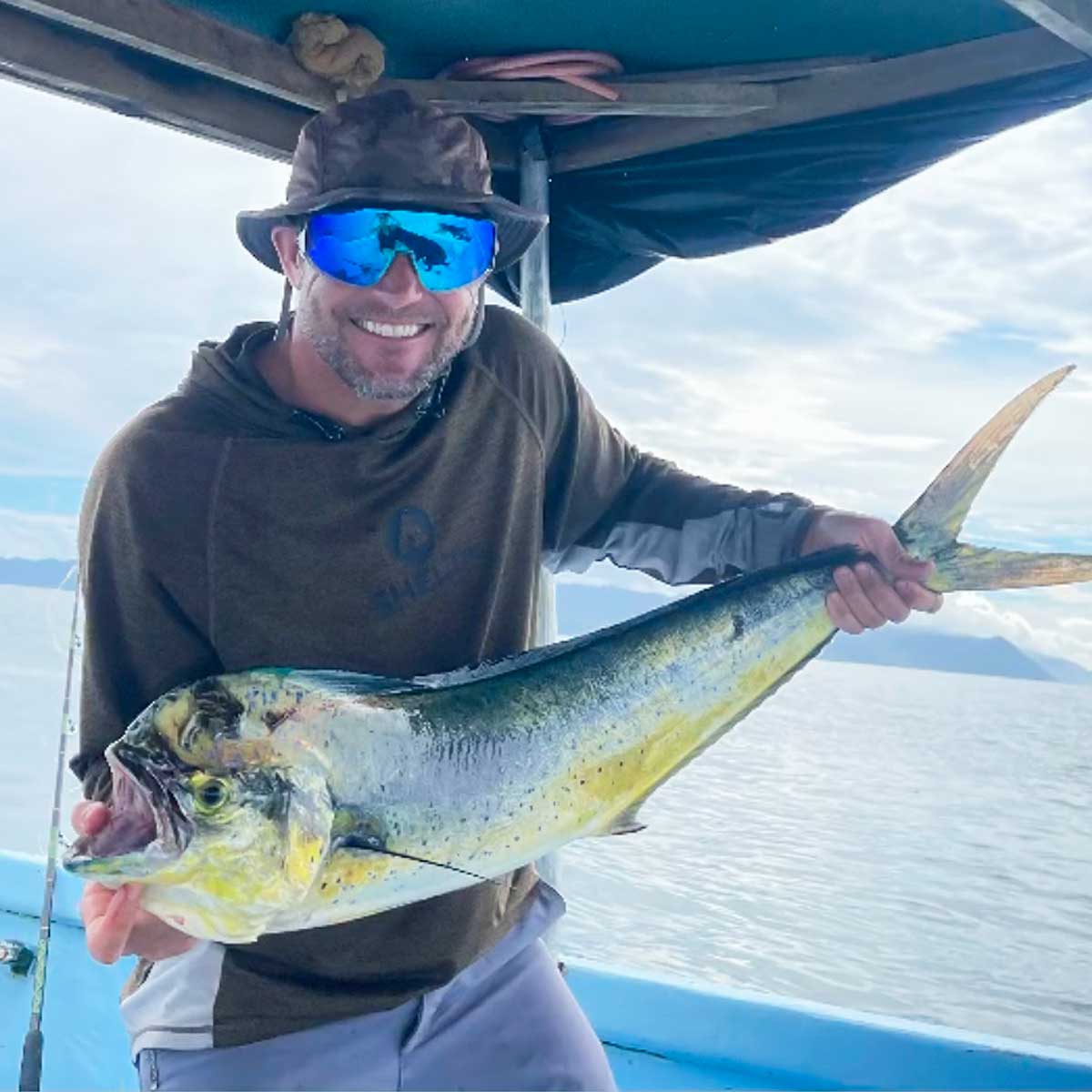 a man holding a dorado he just caught wearing a shelta hat and assault hoodie