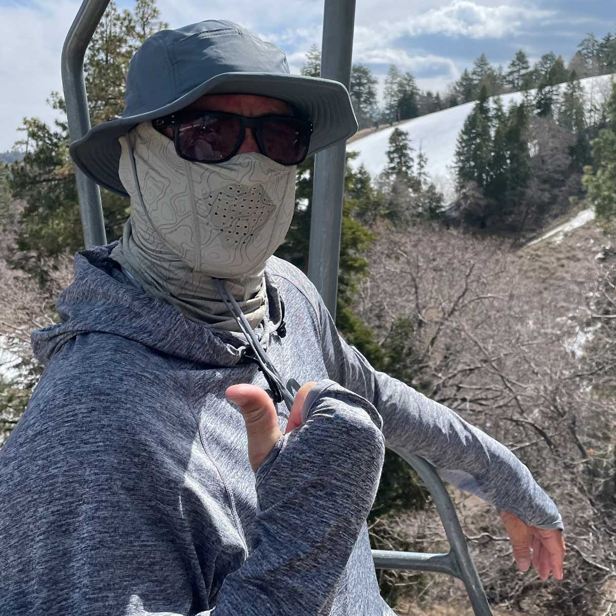 a man sitting on a chairlift giving a thumbs up wearing a shelta sun hat, face gaiter and assault hoodie