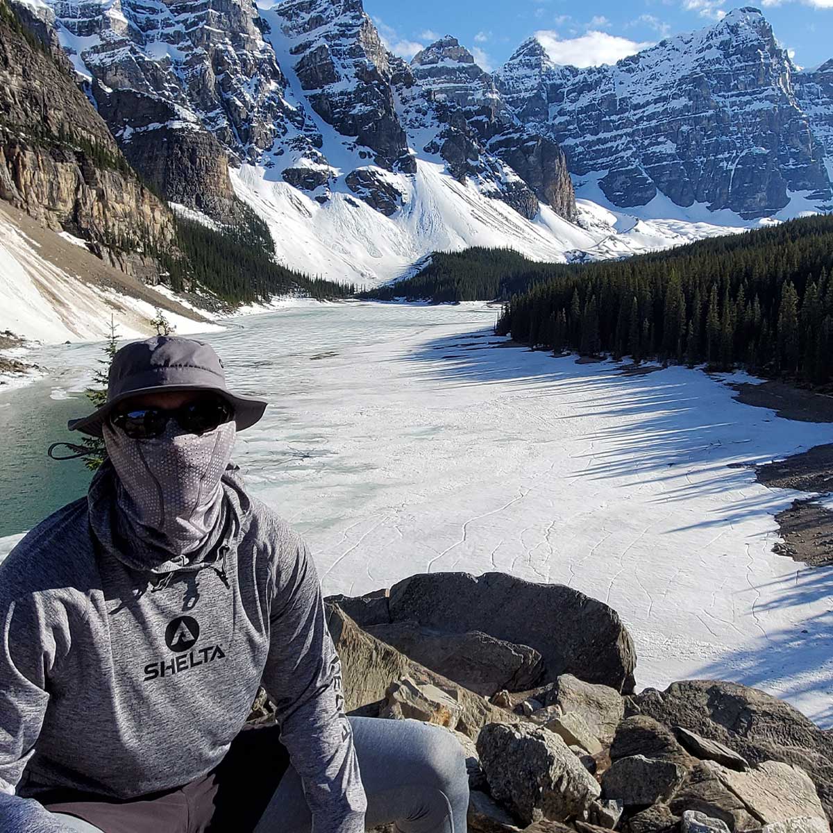 a view of a frozen lake and mountains with a man wearing a shelta assault hoodie, face gaiter and sun hat.