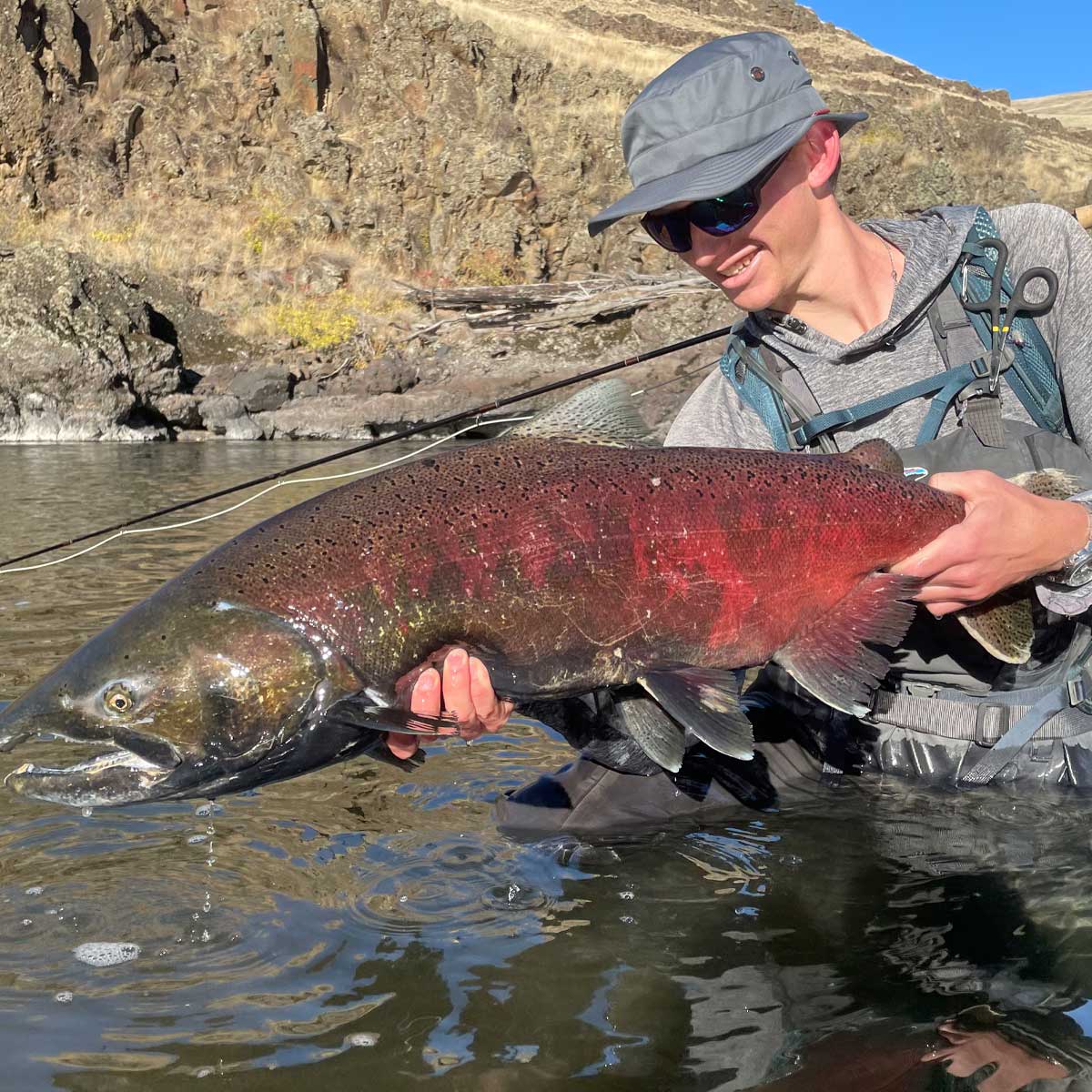 a man standing in a river showing off a huge salmon he just caught wearing a shelta sun hat and assault hoodie