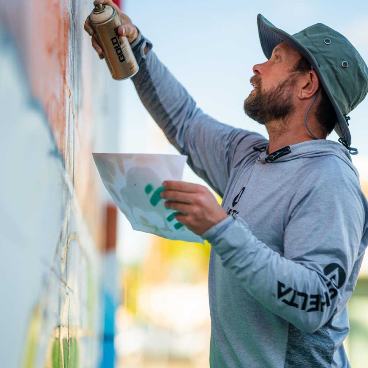 a man spray painting a mural on a building while wearing a shelta sun hat and assault hoodie.
