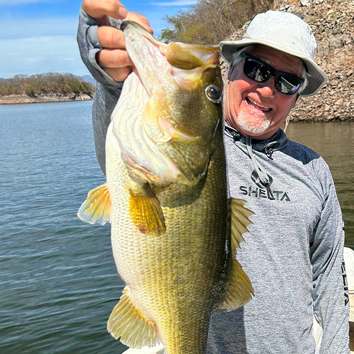 a man holding up a big bass he just caught wearing a shelta sun hat and assault hoodie