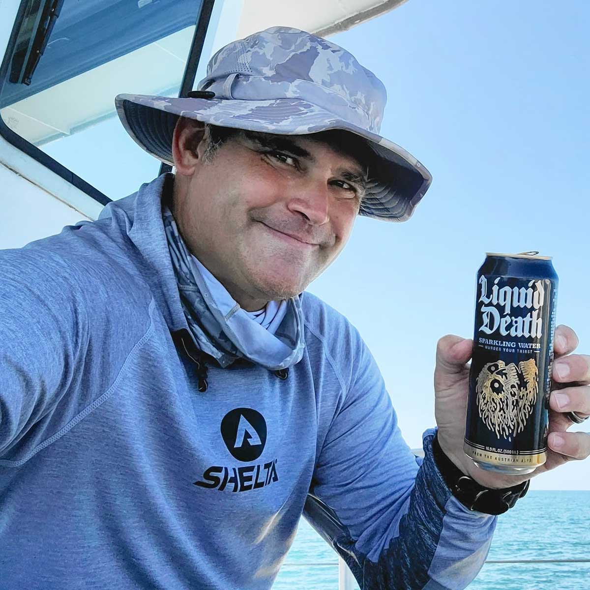 a man on a boat holding up a can of liquid death sparkling water while wearing a shelta sun hat and assault hoodie