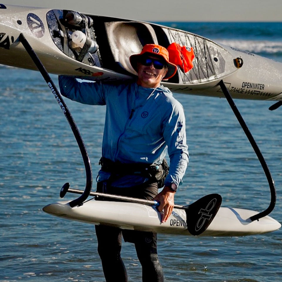 a man carrying an outrigger canoe from the water to the beach wearing a shelta seahawk sun hat