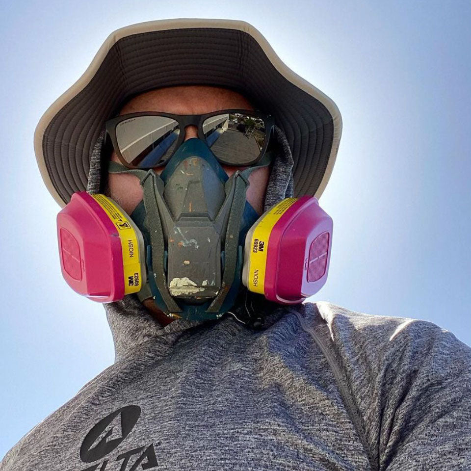 a man wearing a respirator while painting a mural on the side of a building wearing a shelta sun hat
