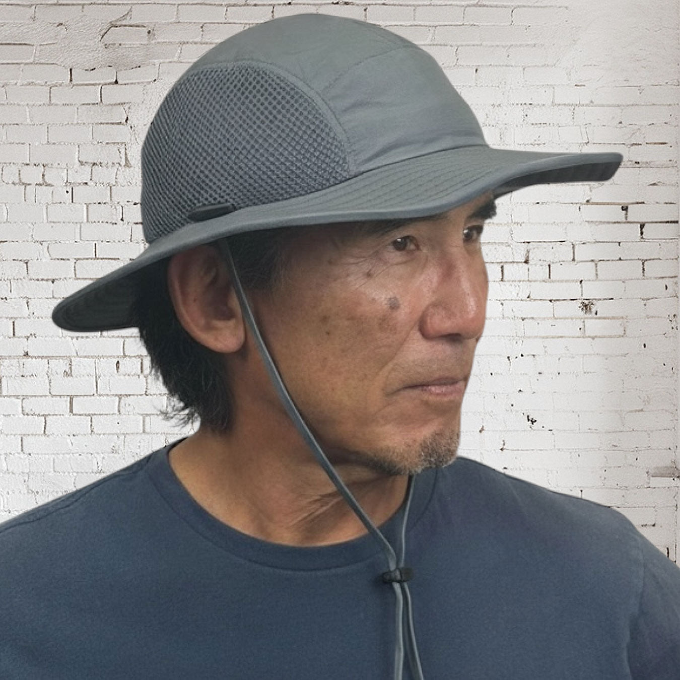Man wearing a gray wide-brimmed hat against a white brick wall.