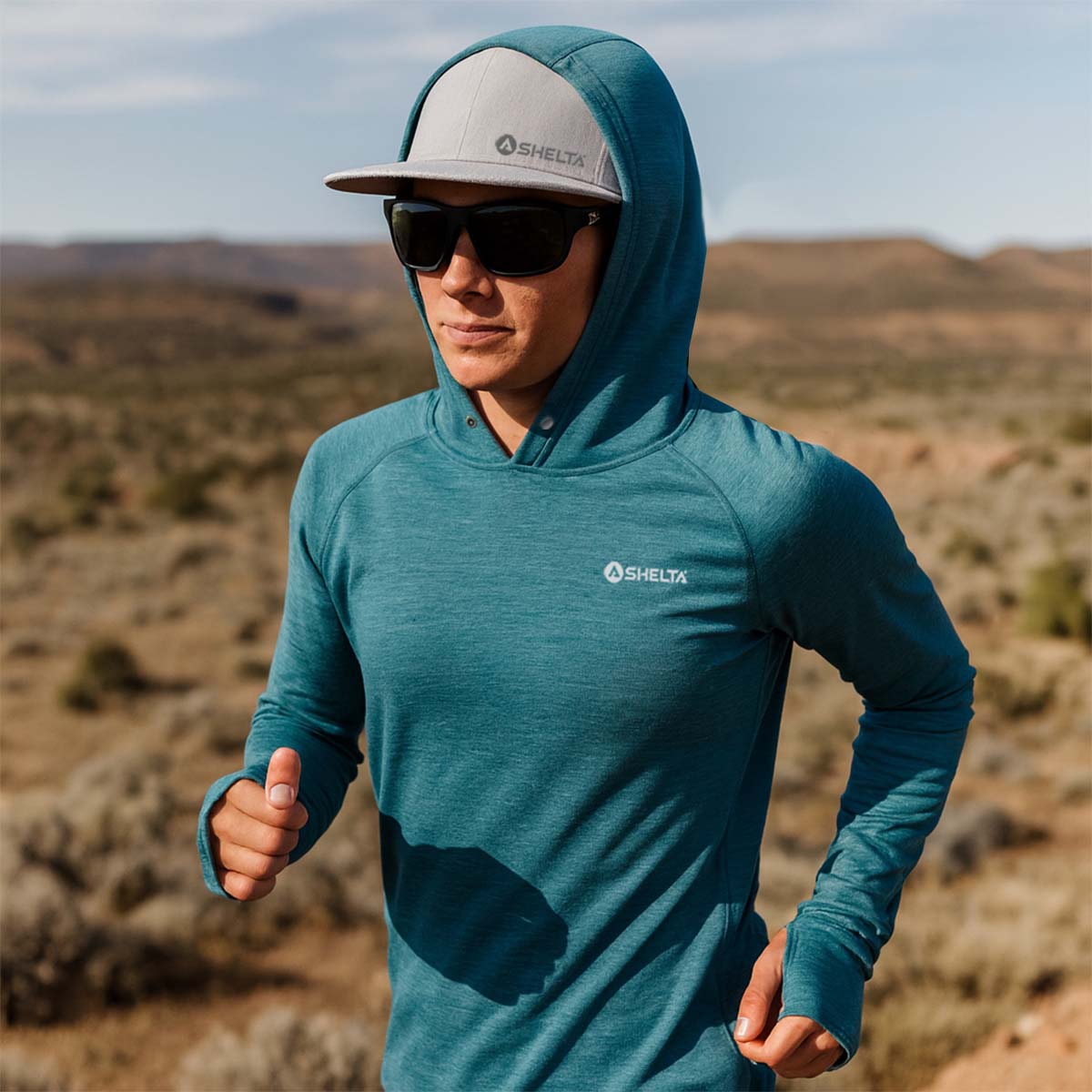 Person wearing a teal hoodie and sunglasses with a desert landscape in the background