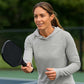 Woman holding a pickleball paddle on a tennis court wearing a gray hoodie with a brand logo.