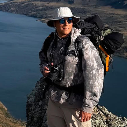 Person with camera and backpack standing on a mountain with a scenic view of water and mountains.