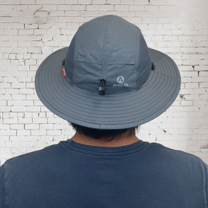 Person wearing a gray wide-brimmed hat with a visible brand logo against a white brick wall.