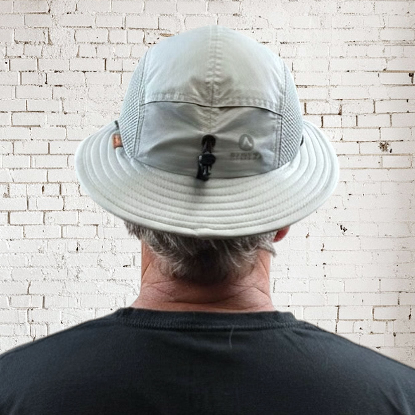Person wearing a light gray bucket hat with a visible brand logo against a white brick wall.