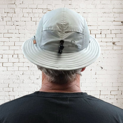 Person wearing a light gray bucket hat with a visible brand logo against a white brick wall.