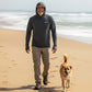 Man walking a dog on a beach wearing a hooded sweatshirt with a brand logo.
