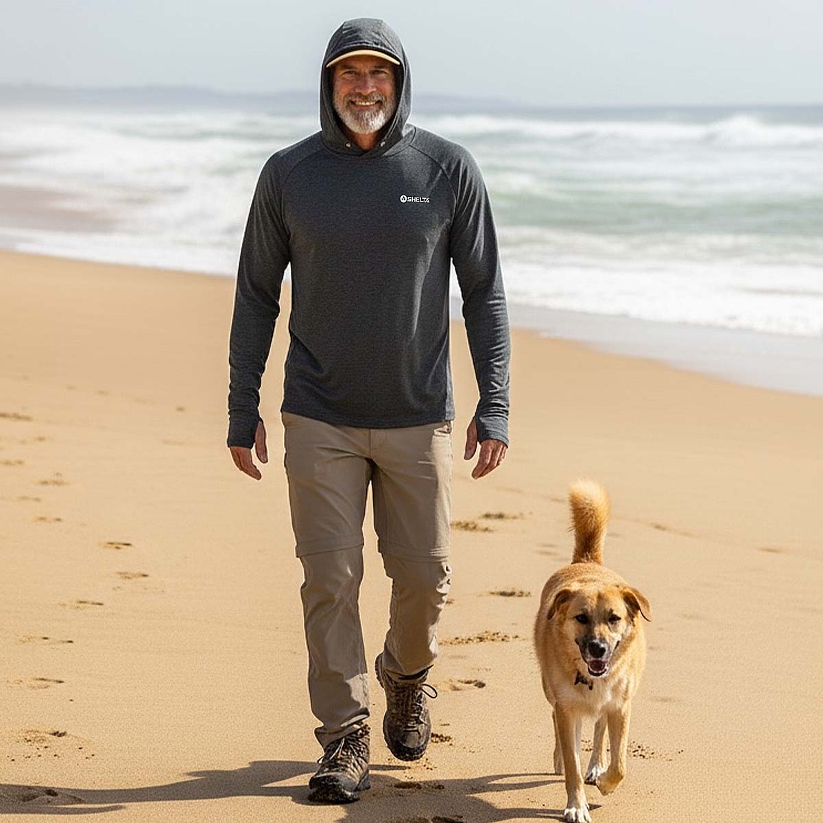 Man walking a dog on a beach wearing a hooded sweatshirt with a brand logo.