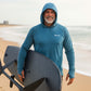 Man in a blue hooded shirt holding a surfboard on a beach