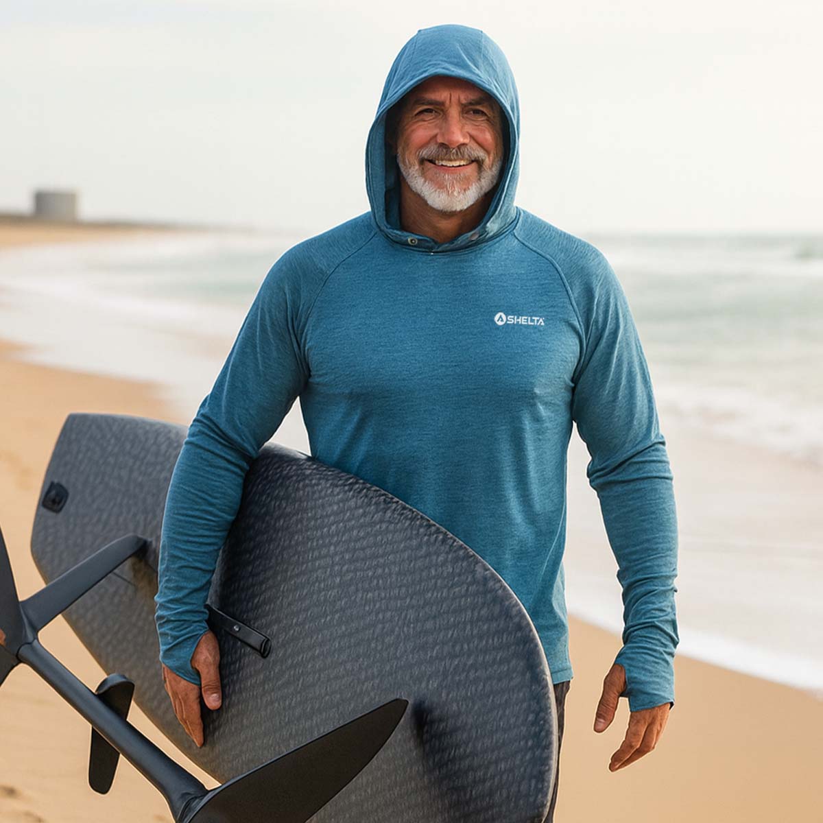 Man in a blue hooded shirt holding a surfboard on a beach
