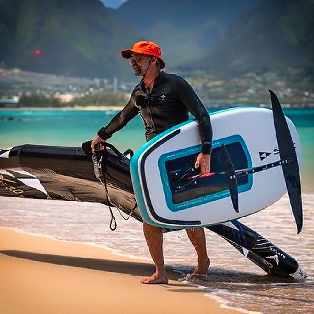 Man carrying a surfboard on a beach with mountains in the background wearing an orange shetla sun hat