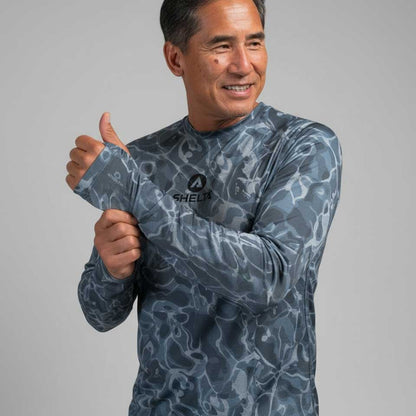 Man wearing a blue camouflage-patterned shirt with a brand logo, giving a thumbs-up gesture on a gray background.