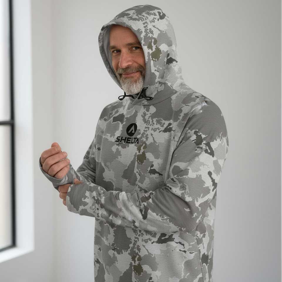 Man wearing a camouflage hoodie with a shelta logo on a neutral background