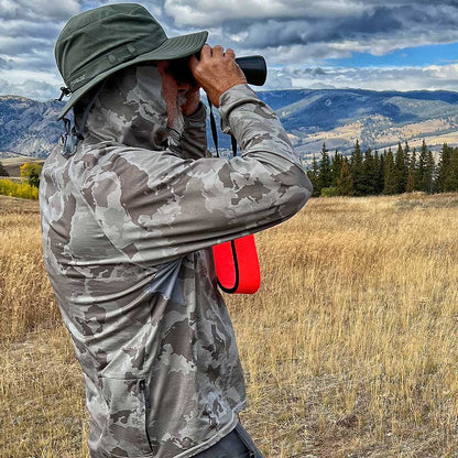Person in camouflage clothing using binoculars in a scenic outdoor setting with mountains and trees.