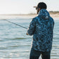 Man fishing by a body of water wearing a blue camouflage jacket.