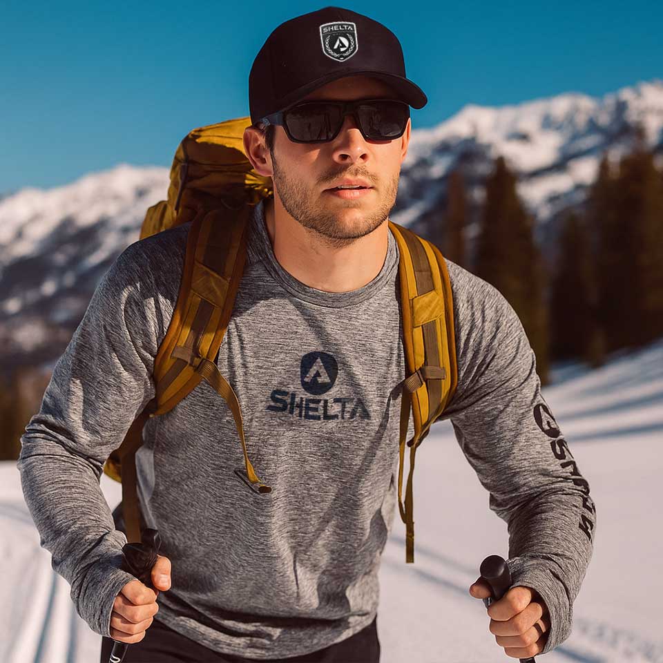 Man hiking in snowy mountains wearing a gray Shelta shirt and black cap.