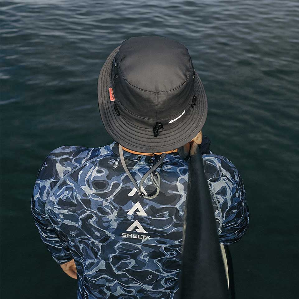 Person wearing a Shelta Sun hat and blue camouflage jacket with 'Shelta' branding, standing by water.