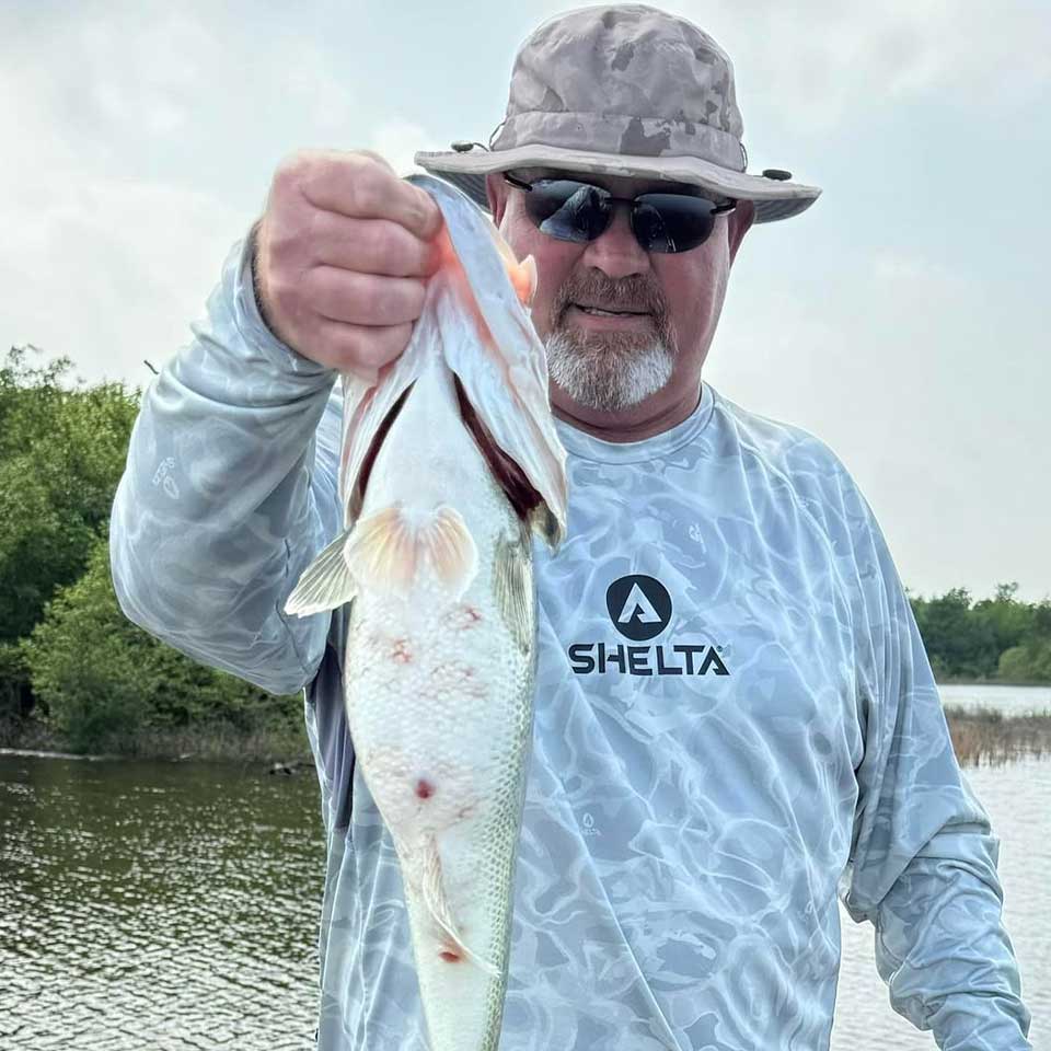 Man holding a fish by a body of water, wearing a Selta shirt.
