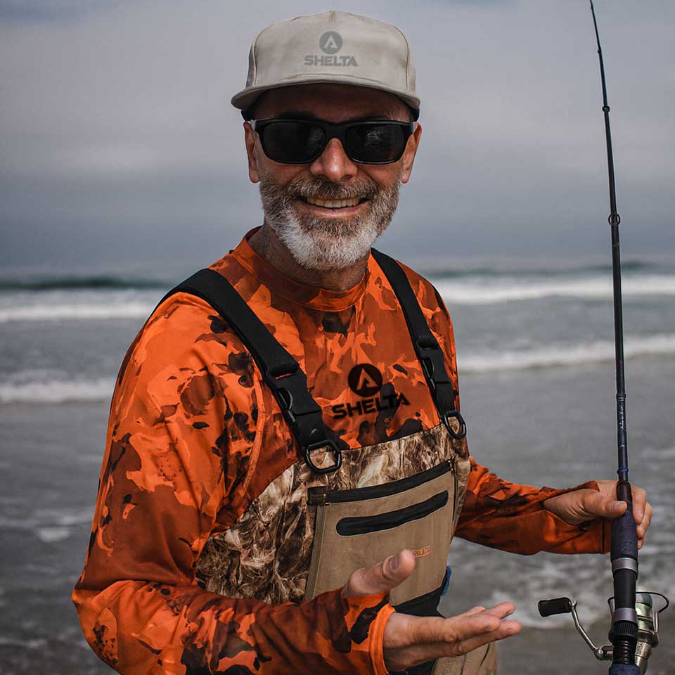 Man in orange camouflage shirt and beige cap holding a fishing rod on a beach.