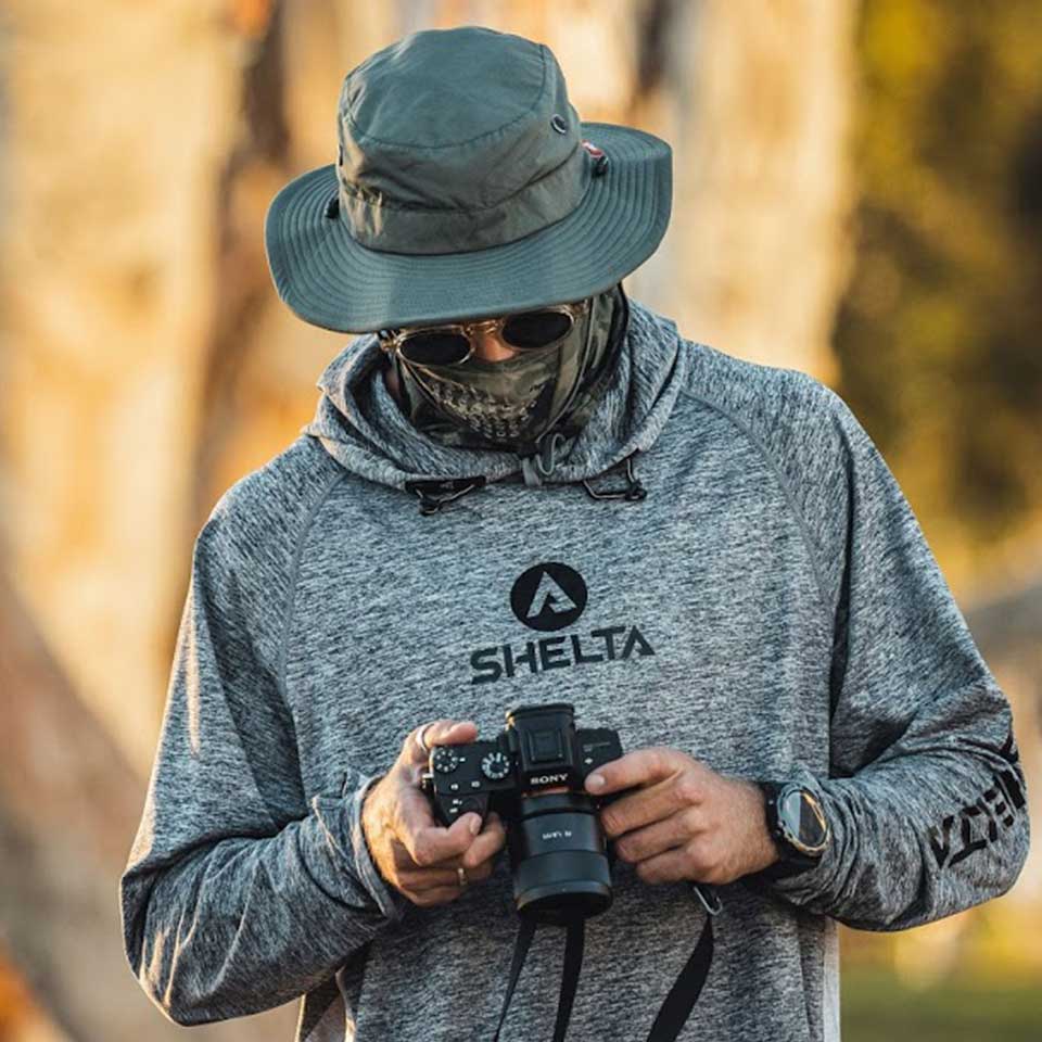 Person wearing a gray hoodie with 'SHELTA' logo, holding a camera in a forest setting