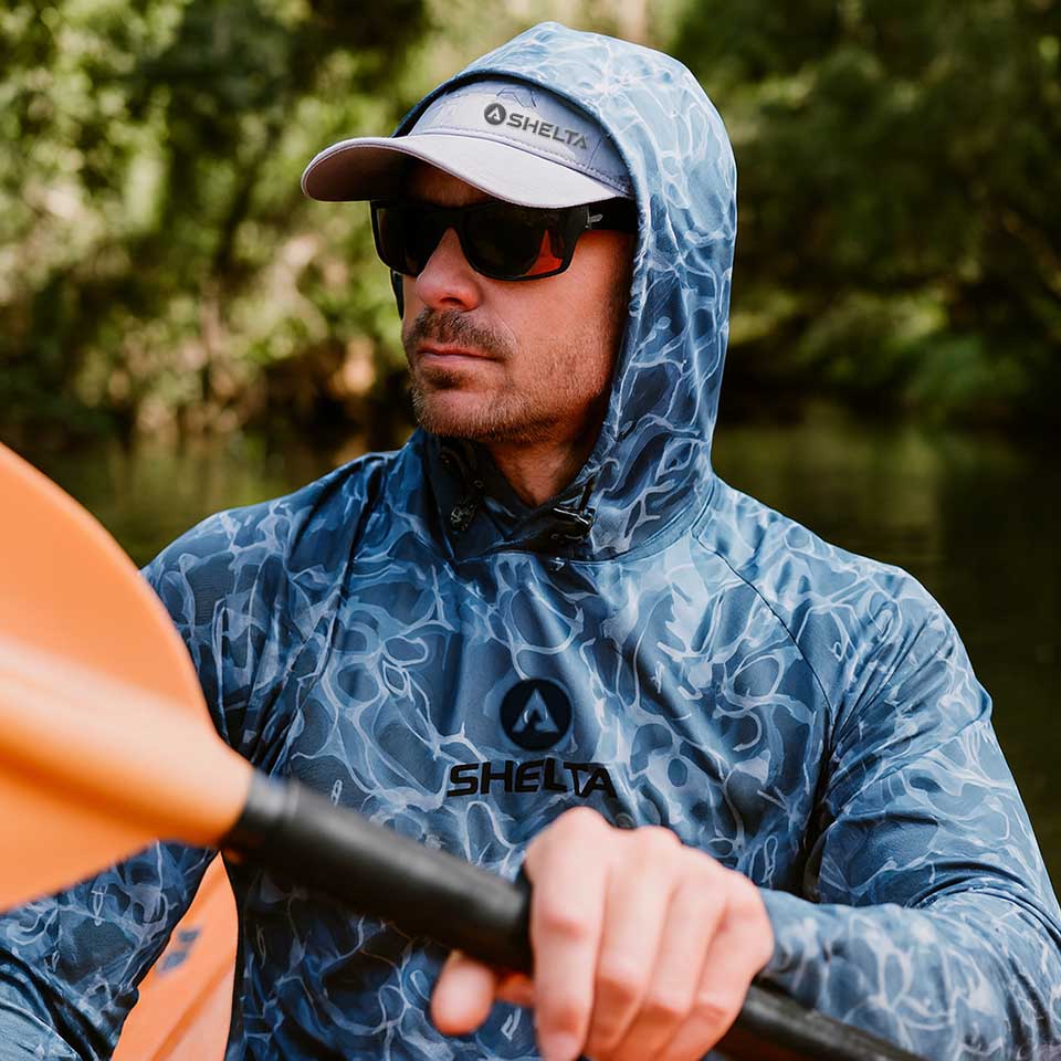Man in a blue hooded jacket with Shelta logo holding a paddle, surrounded by greenery.