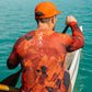 Man paddling a kayak in clear blue water wearing an orange and black patterned shirt.