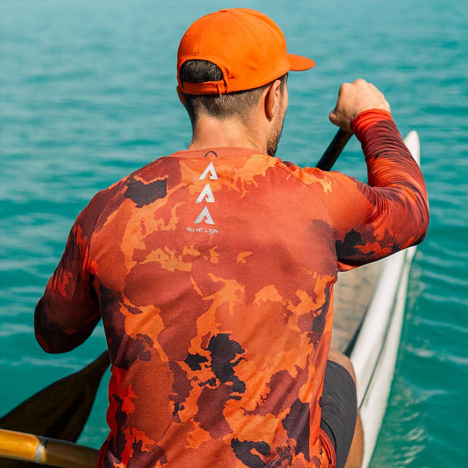 Man paddling a kayak in clear blue water wearing an orange and black patterned shirt.