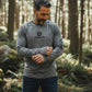 Man wearing a gray long-sleeve shirt with 'Shelta' logo in a forest setting