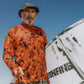 Man wearing an orange camouflage shirt with a logo, holding a  surfboard against a blue sky.