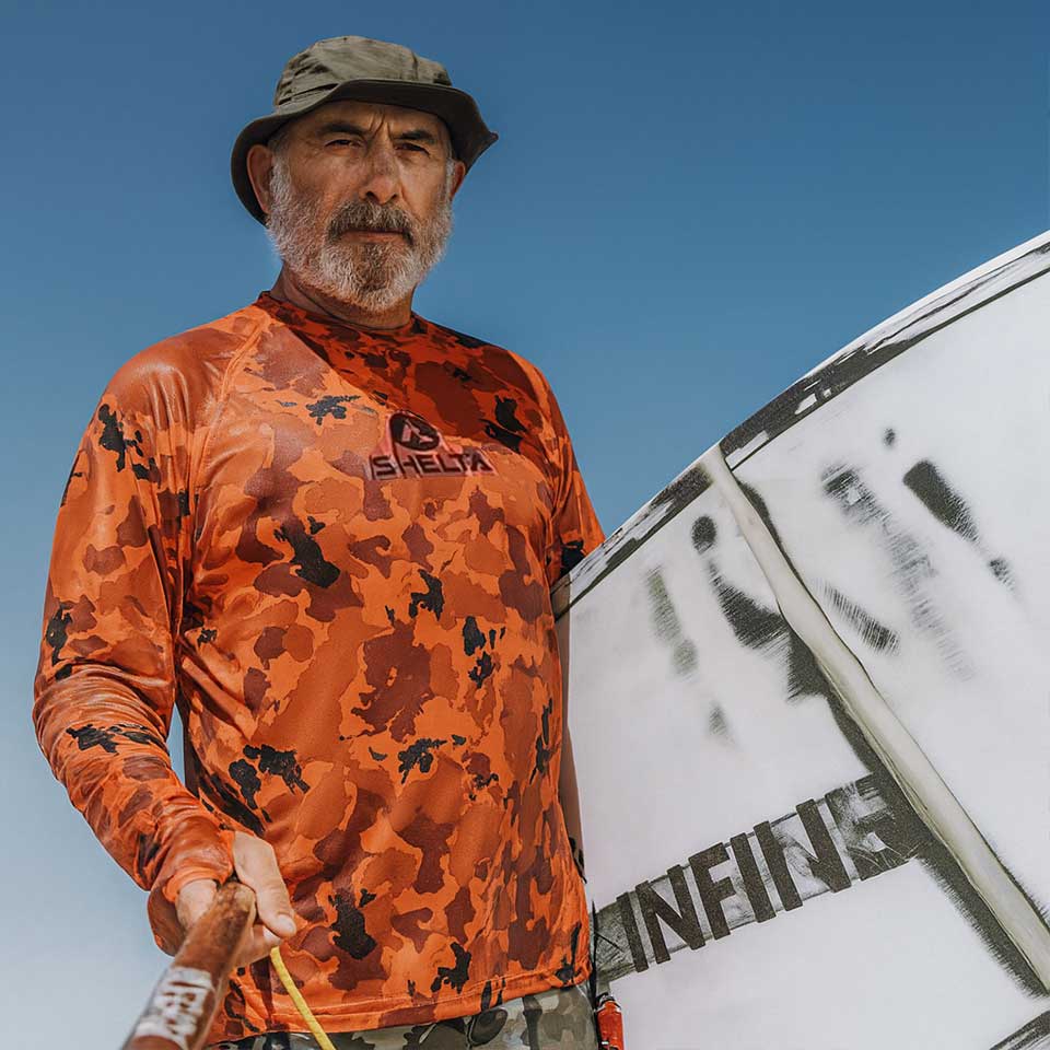 Man wearing an orange camouflage shirt with a logo, holding a  surfboard against a blue sky.