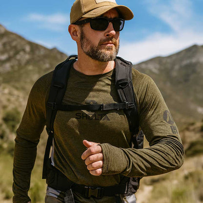 Man in outdoor setting wearing a green long-sleeve shirt, cap, and sunglasses with mountains in the background.