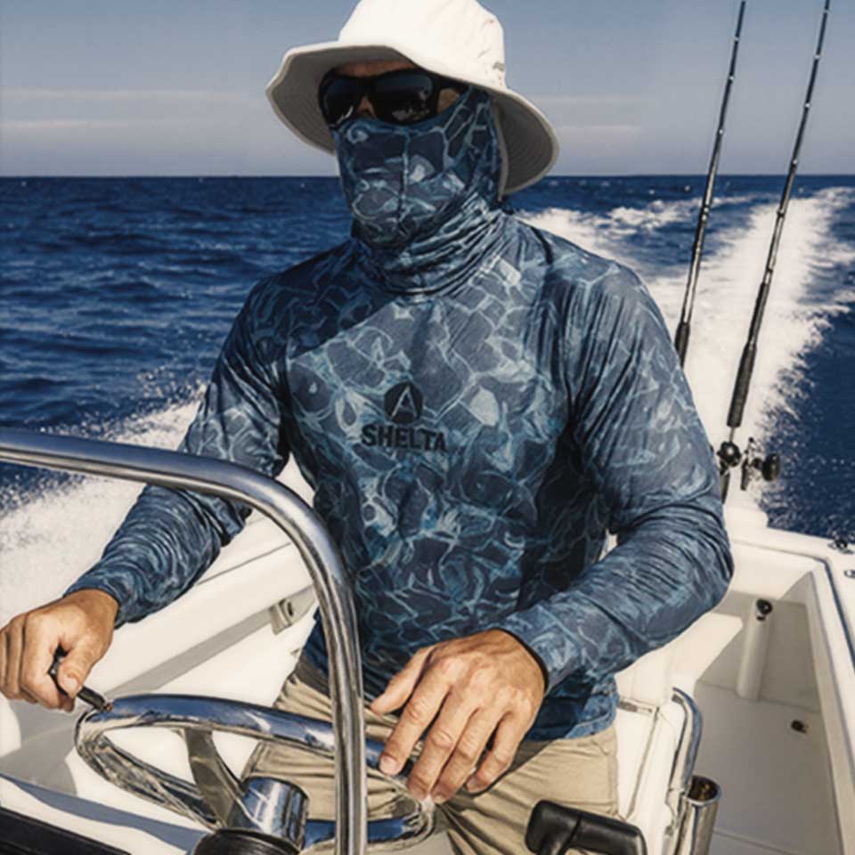 Person on a boat wearing a blue camouflage shirt with a brand logo, sitting at the helm.