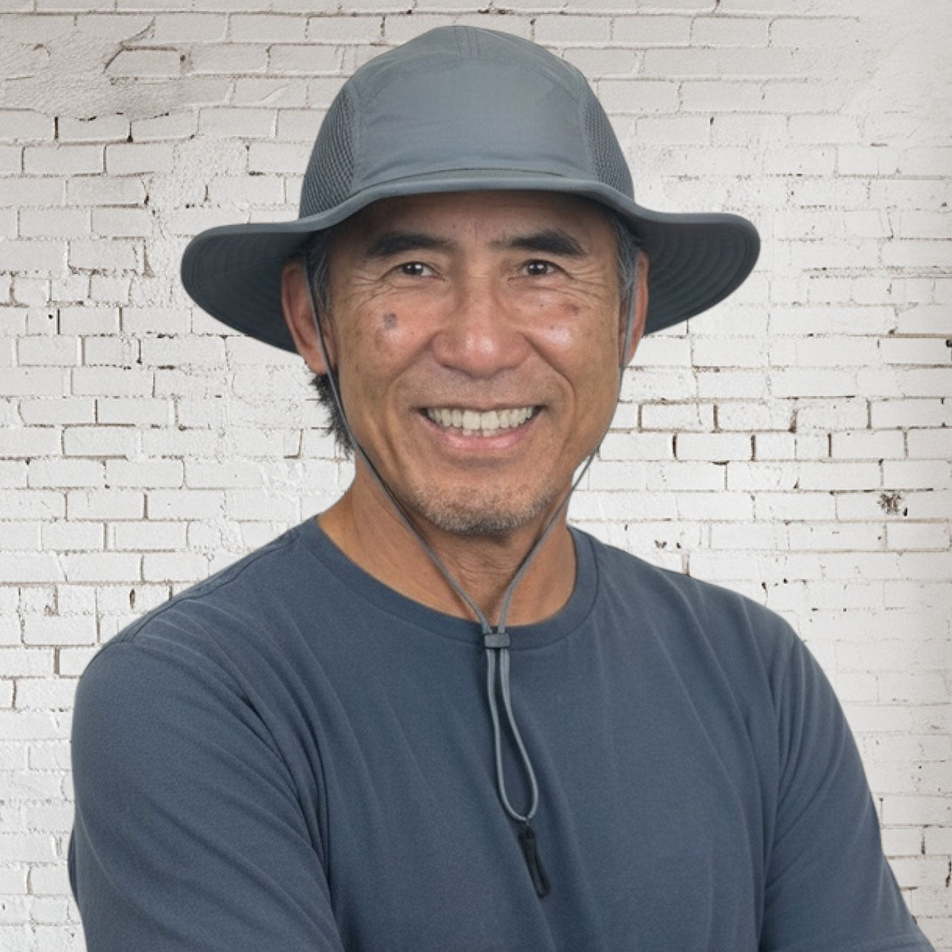 Man wearing a gray bucket hat and matching shirt against a white brick wall.