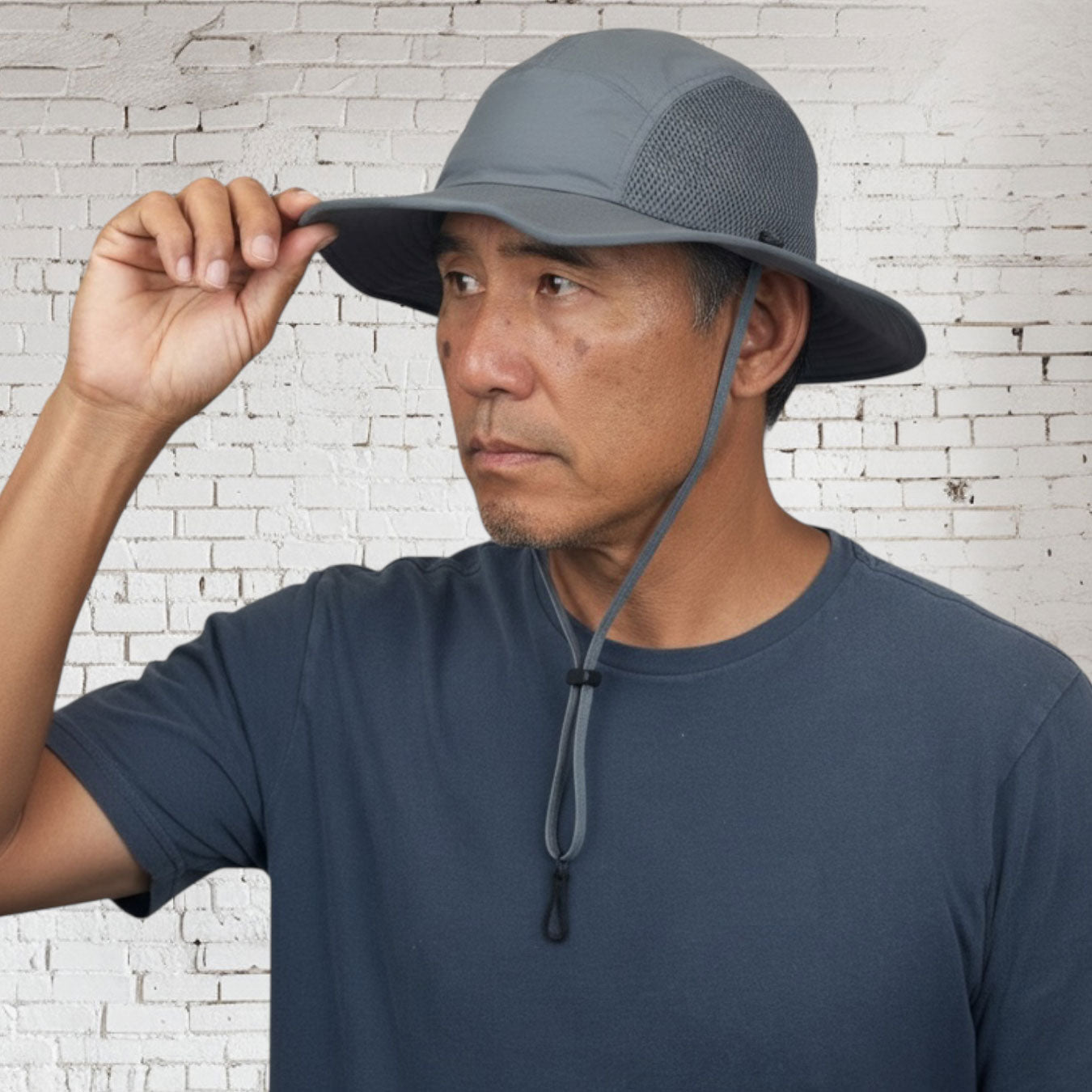 Man wearing a gray bucket hat against a white brick wall.