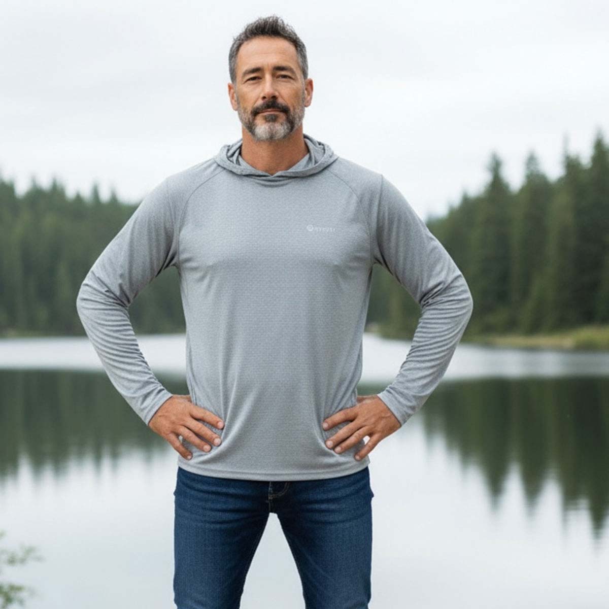 Man wearing a gray hoodie and blue jeans standing by a lake with trees in the background
