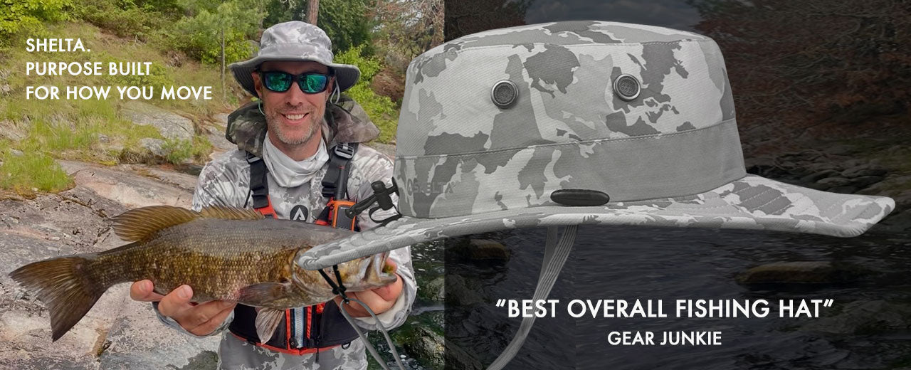 A man wearing a camouflage fishing hat and sunglasses holds a fish while standing outdoors by a river, with a large close-up of the hat on the right and text reading 'SHELTA. PURPOSE BUILT FOR HOW YOU MOVE' and 'BEST OVERALL FISHING HAT GEAR JUNKIE'.