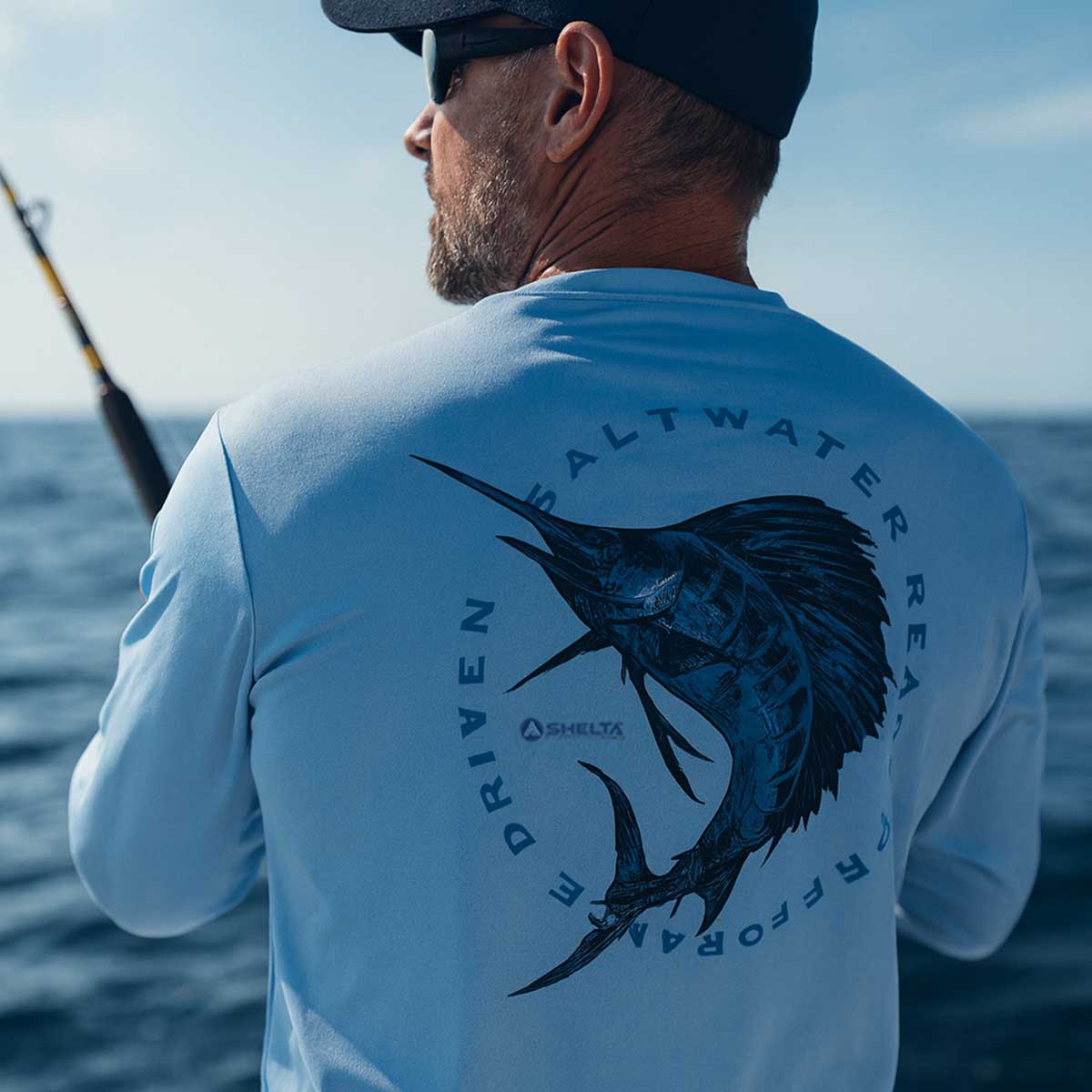 Man on a boat wearing a light blue shirt with a fish graphic and text, looking out over water.