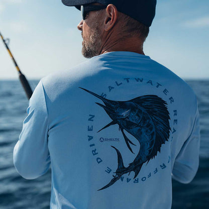 Man on a boat wearing a light blue shirt with a fish graphic and text, looking out over water.
