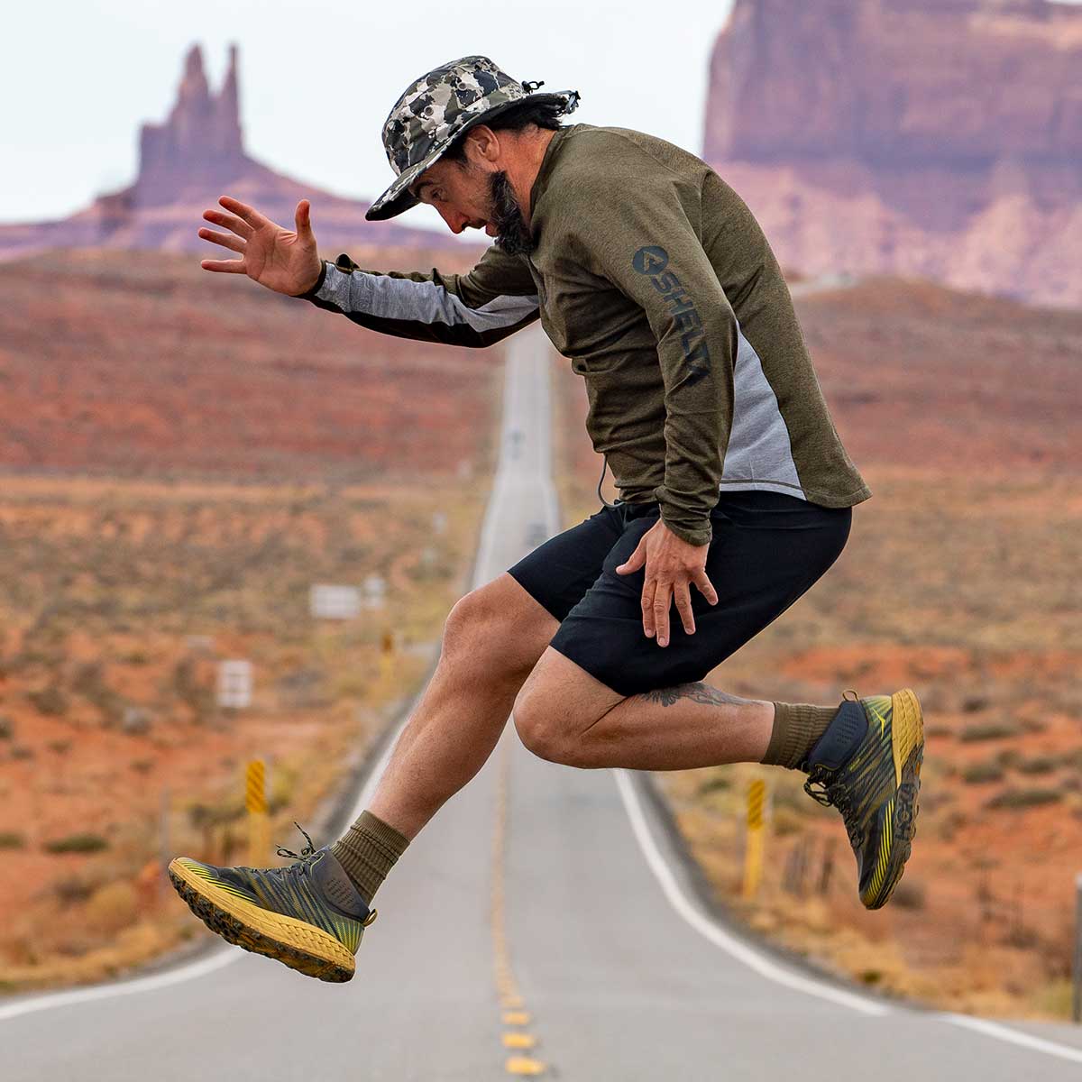 a man jumping in the air in the middle of the desert wearing a shelta sun hat and aquaterra sun shirt