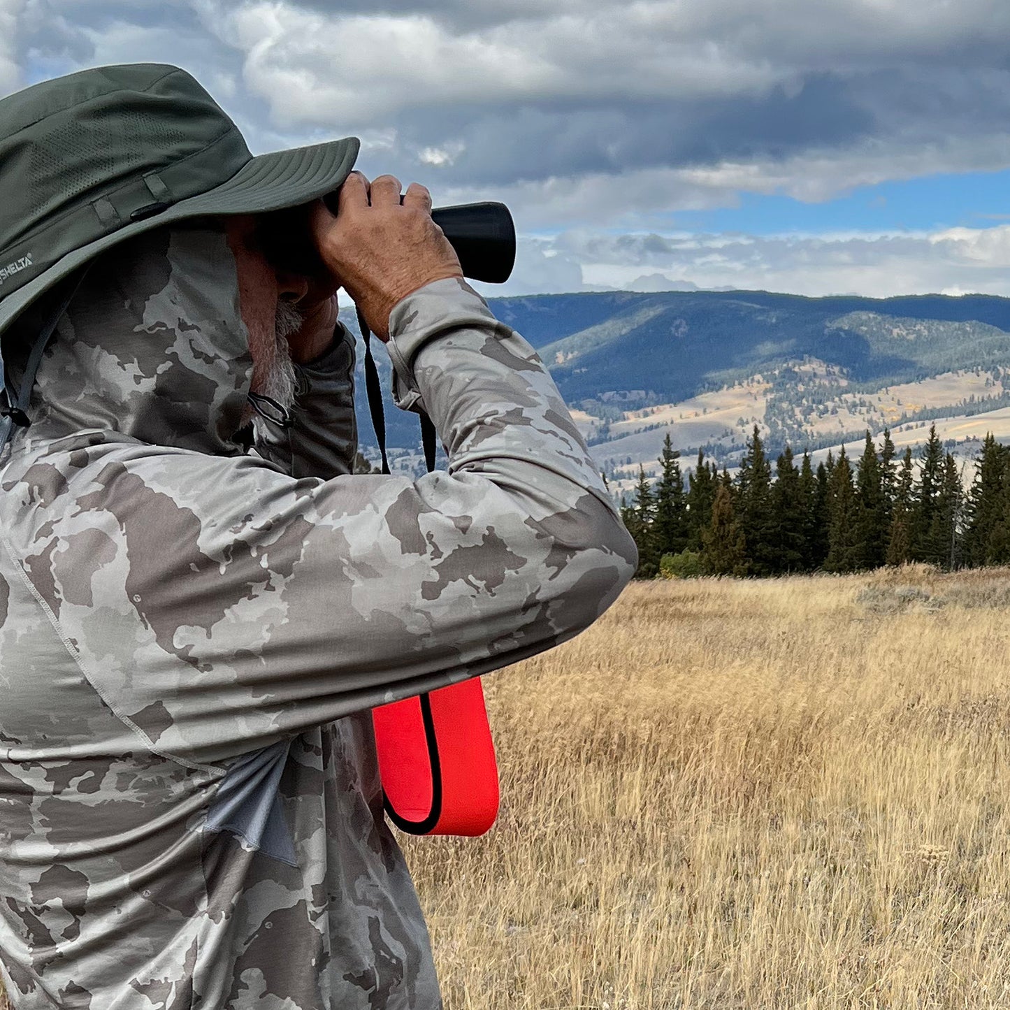 Person in camouflage clothing using binoculars in a scenic outdoor setting with mountains and trees.